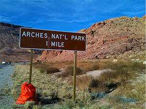 Photo of roadway approaching Arches Natoinal Park. The area is litter free. An orange trash bag leans against the sign that says "Arches Nat'l Park 1 mile"